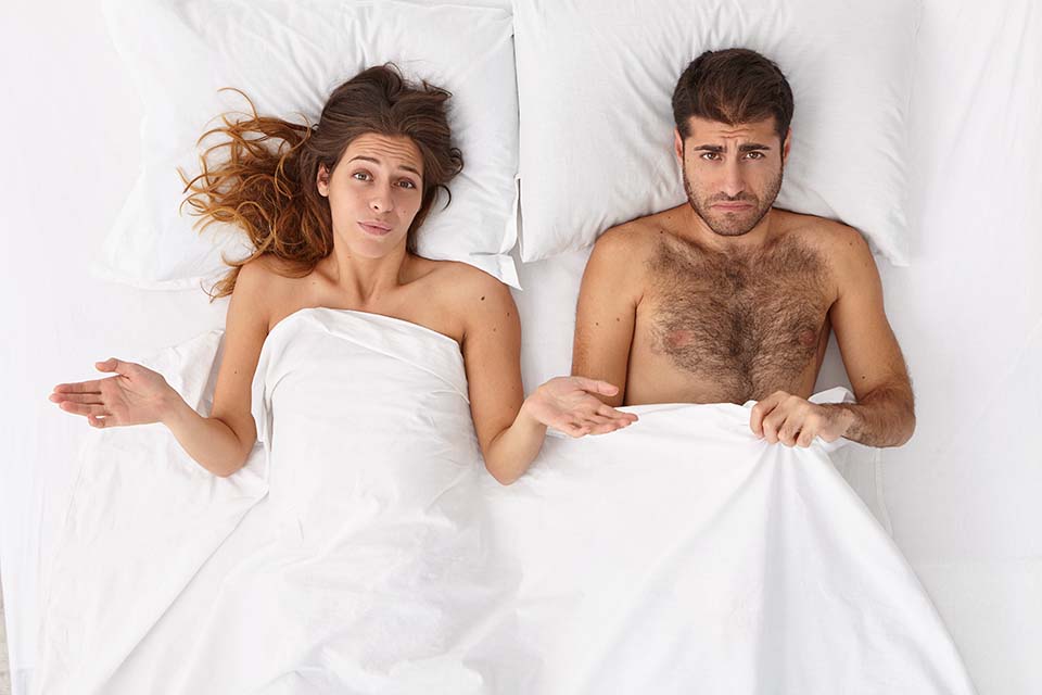 a couple are under the bed sheets. The woman has a questioning expression and the man has a worried expression